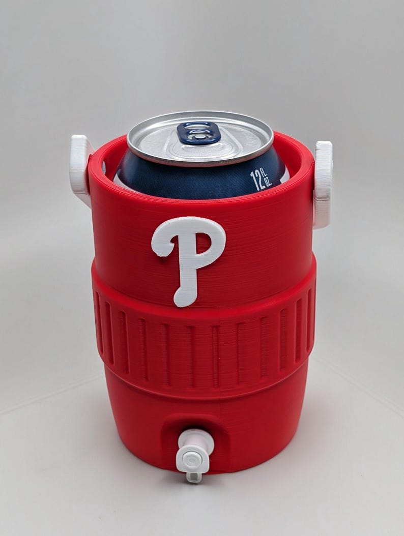 May include: A red cylindrical beverage cooler designed to hold a 12 fl oz can, featuring a white letter P logo on the front, two white side handles, and a small white spigot at the base. The cooler has a ribbed texture around the middle section.