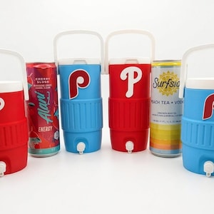 May include: Several beverage coolers in red and blue with white handles and spouts, each with a white "P" logo. Cans of soda and energy drinks are next to the coolers. The cans include "Flight", "Alani Energy", "Surfside", and "Diet Coke".