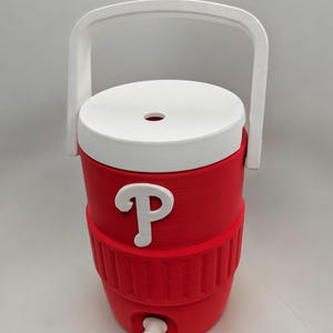 May include: A red beverage cooler with a white lid, a white handle, and a white spigot at the base. The front of the cooler features a white letter P logo. The cylindrical container has a textured ribbed section around the middle and a circular opening in the center of the lid.
