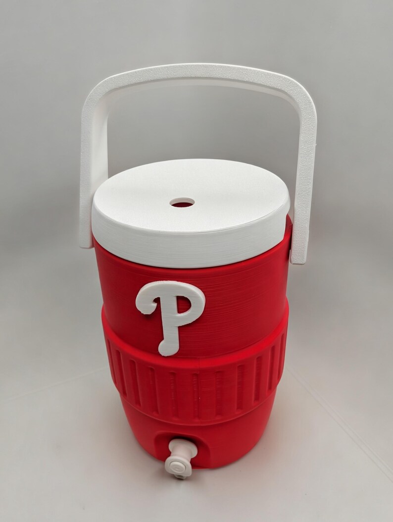 May include: A red beverage cooler with a white lid, a white handle, and a white spigot at the base. The front of the cooler features a white letter P logo. The cylindrical container has a textured ribbed section around the middle and a circular opening in the center of the lid.