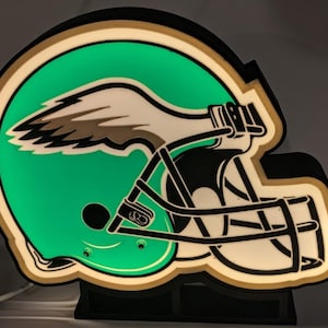 May include: A lighted sign in the shape of a football helmet, featuring a green background with a white and black wing design. The helmet has a black facemask and is outlined in gold and white.