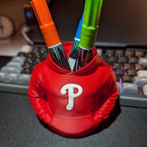 May include: A red pen holder shaped like a hoodie, with a white "P" logo on the front. Two markers, one orange and one green, are visible inside. The pen holder is on a black surface, with a keyboard in the background.