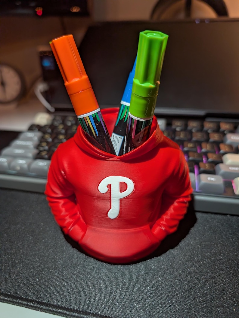 May include: A red pen holder shaped like a hoodie, with a white "P" logo on the front. Two markers, one orange and one green, are visible inside. The pen holder is on a black surface, with a keyboard in the background.