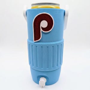 May include: A light blue beverage cooler with a white handle and a white spigot at the base. The front features a large, maroon and white letter P logo with a textured, glittery finish. A yellow and silver beverage can is inserted at the top.