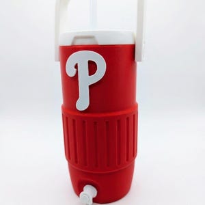 May include: A red insulated beverage cooler with a white handle and a white lid. The cooler features a prominent white letter P logo on the front and a small white spigot at the base. A clear plastic straw extends from the top of the lid.