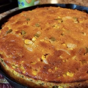 May include: A golden-brown cornbread, baked in a black cast iron skillet. The cornbread contains kernels of corn, diced green peppers, and onions. The edges are crispy, and the top is slightly browned.