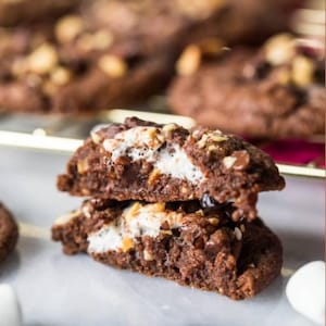 Puede incluir: Dos galletas de chocolate cortadas por la mitad, revelando un centro de malvavisco. Las galletas están cubiertas con nueces y chispas de chocolate. Se ven más galletas en el fondo.