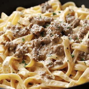 Könnte beinhalten: Nahaufnahme eines cremigen Pastagerichts mit Fettuccine-Nudeln, belegt mit zerbröselter Wurst und geriebenem Käse. Das Gericht ist mit gehackter Petersilie garniert und wird in einer schwarzen Schüssel serviert.