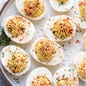 Può includere: Un piatto di uova alla diavola, ciascuna dimezzata e riempita con un ripieno giallo. Guarnite con paprika e aneto fresco, le uova sono disposte su un piatto bianco con un bordo marrone, perfette per una festa o un antipasto.