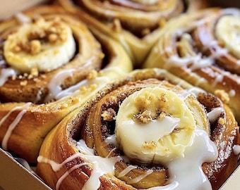 Peut inclure: Une boîte de brioches à la cannelle fraîchement cuites, garnies de tranches de banane, de glaçage blanc et de noix hachées. Les brioches sont dorées et disposées dans une boîte en carton. Le glaçage est versé sur les brioches.
