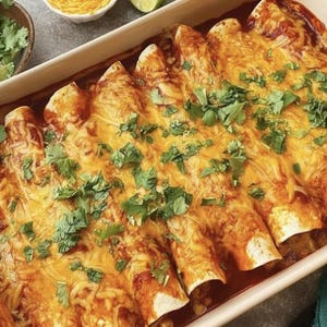 A baked enchilada casserole dish filled with a rich, cheesy sauce and topped with fresh cilantro and other green herbs. The dish is presented in a white baking dish, and there are a few pieces of cilantro sprinkled on top.