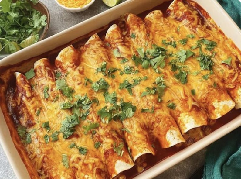 A baked enchilada casserole dish filled with a rich, cheesy sauce and topped with fresh cilantro and other green herbs. The dish is presented in a white baking dish, and there are a few pieces of cilantro sprinkled on top.