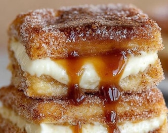 Op de afbeelding: Een stapel van twee vierkante gebakjes met een romige witte vulling en een drizzle van karamelsaus. De gebakjes zijn bedekt met kaneelsuiker, wat zorgt voor een goudbruine, getextureerde uitstraling. De karamelsaus druipt langs de zijkanten.