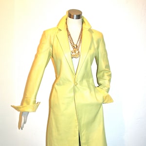 May include: A long, light yellow leather jacket with a single button closure. The jacket has a collar and long sleeves with a satin lining.