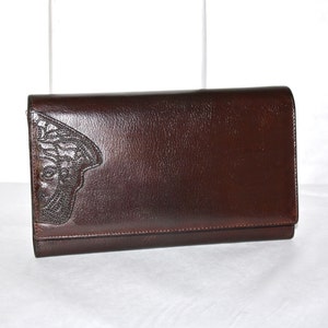 May include: A dark brown leather clutch with a flap closure. The clutch features a decorative embossed design on the front left corner. The leather has a slight sheen, and the overall design is rectangular.