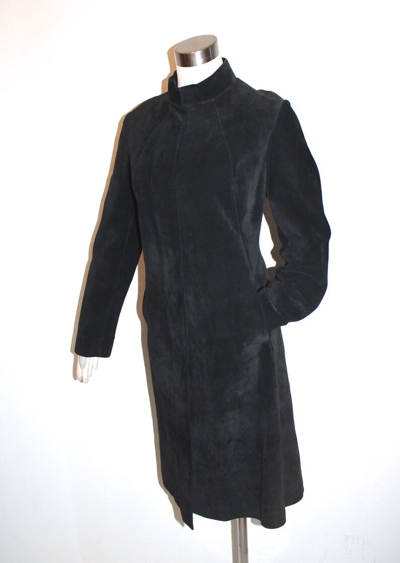 May include: A black suede coat with a high collar and long sleeves. The coat has a tailored fit and a knee-length hem. The coat is displayed on a mannequin against a white background. The coat has two pockets.