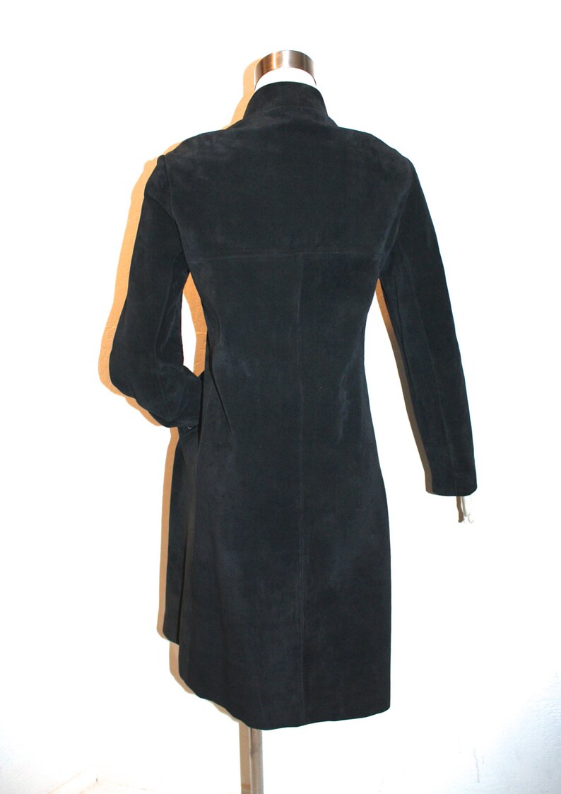 May include: A black suede coat with a high collar and long sleeves. The coat is knee-length and has a tailored fit. The back of the coat is visible, showing the seams and the overall shape. The coat is displayed on a mannequin.