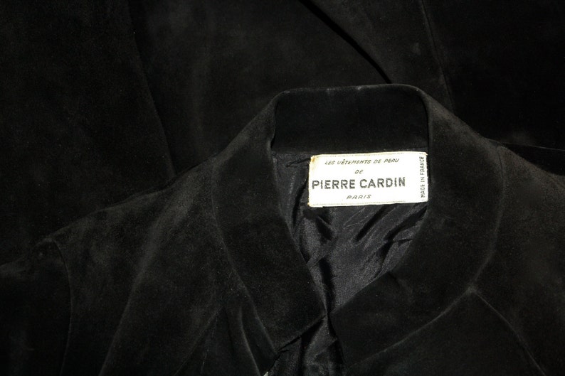 May include: Close-up of a black suede jacket with a notched collar. The label reads "LES VETEMENTS DE PEAU DE PIERRE CARDIN PARIS". The jacket's interior lining is visible.