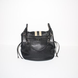 Pu&ograve; includere: Borsa a mano in pelle nera con dettaglio a strisce beige e bianche sulla patta. La borsa presenta finiture color argento, un manico superiore e chiusure con coulisse. Un piccolo logo argentato &egrave; visibile sul davanti.