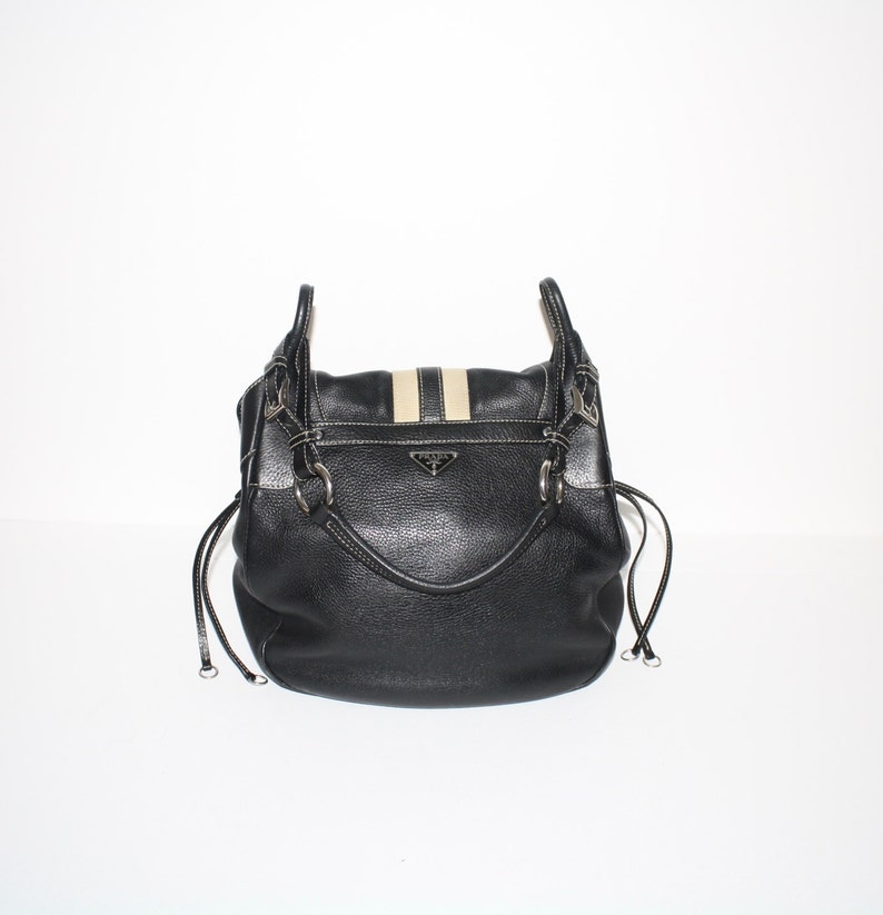 Pu&ograve; includere: Borsa a mano in pelle nera con dettaglio a strisce beige e bianche sulla patta. La borsa presenta finiture color argento, un manico superiore e chiusure con coulisse. Un piccolo logo argentato &egrave; visibile sul davanti.