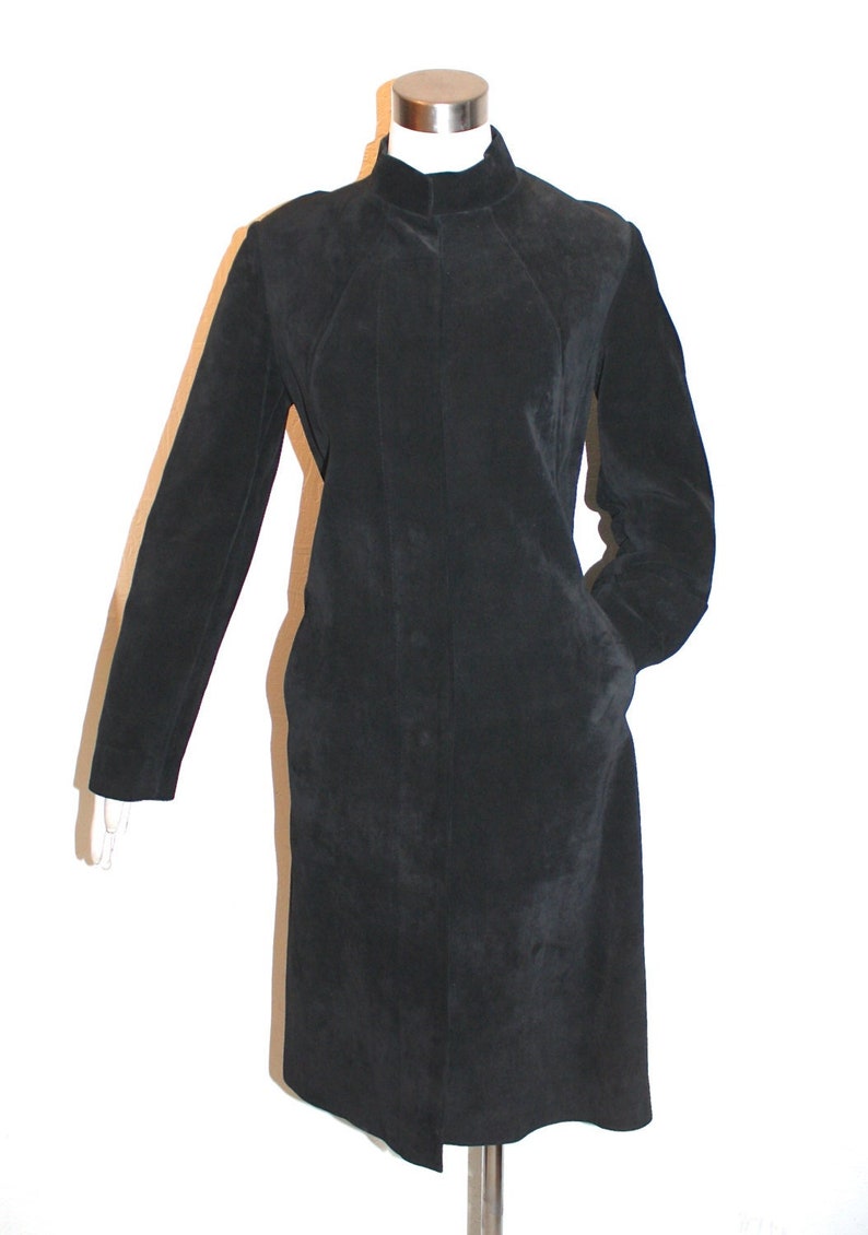 May include: A black suede coat with a high collar and long sleeves. The coat has a button-down front and two pockets. The coat is displayed on a mannequin against a white background. The coat is knee-length.