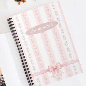Puede incluir: Un cuaderno de espiral con una cubierta a rayas rosas y blancas, adornada con motivos florales y un lazo rosa. El nombre "Samantha" está escrito en la portada. El cuaderno está abierto, mostrando páginas en blanco.
