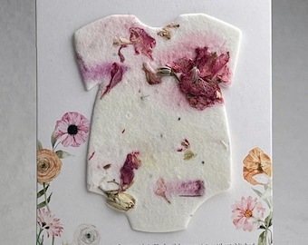 May include: A white card featuring the text Celebrating Charlie! and a onesie-shaped piece of seed paper embedded with dried pink flower petals. The card includes floral illustrations and instructions to cover with 1/8 inch of soil.
