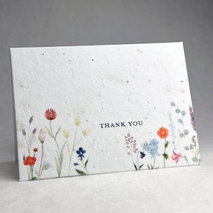 May include: A white rectangular folded card with a textured, speckled surface. The front features a watercolour illustration of various wildflowers in red, orange, blue, and green along the bottom edge, with the text THANK YOU printed in black.