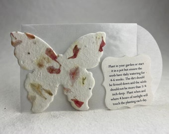 May include: A white butterfly-shaped seed paper with pink and brown flower petals embedded in it. The paper is attached to a white card with instructions on how to plant the paper: "Plant in your garden or start it in a pot but ensure the seeds have daily watering for 4-6 weeks. The dirt should be firmed down and the seeds should not be more than 1/4 inch deep. Plant when and where 4 hours of sunlight will touch the planting each day."