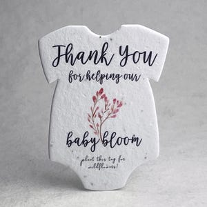 May include: A white, textured, onesie-shaped paper tag featuring black text that reads Thank You for helping our baby bloom, a small illustration of pink floral sprigs, and the text plant this tag for wildflowers at the bottom.