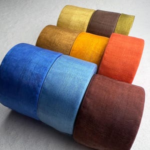 May include: A collection of nine cylindrical spools of velvet ribbon arranged in three rows. The colors include shades of royal blue, light blue, dark brown, mustard yellow, golden brown, bright orange, olive green, and deep chocolate brown.