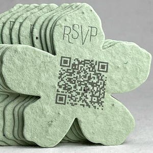 May include: A stack of light green, flower-shaped cards. Each card has a QR code and the word "RSVP" printed on it. The cards are made of a textured, possibly recycled, paper material.