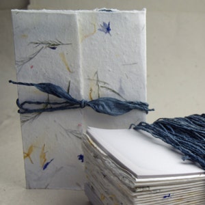 May include: Two handmade journals with blue string covers. The journal on the left is wrapped in a white paper with blue and yellow flowers. The journal on the right is wrapped in a white paper with blue string.