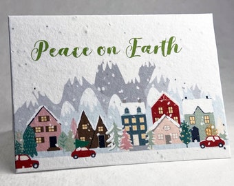 May include: A Christmas card with a winter village scene. The card is white with green text that reads "Peace on Earth". The village is made up of small houses with snow-covered roofs and red cars driving down the street. There are trees and mountains in the background.