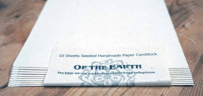 May include: A stack of 10 sheets of seeded handmade paper cardstock. The paper is white and has a natural texture. The packaging has a black and white design with the text "OF THE EARTH" and the words "The bags we use are biodegradable bopp cellophane".