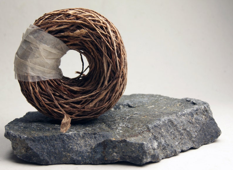 May include: A spool of brown twine wrapped in a white paper band, sitting on a gray rock.