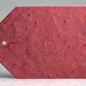 May include: A textured, tag-shaped label in a deep red hue. The tag has a small, circular hole near the top and a pointed bottom edge. The surface has a speckled appearance, suggesting it's made from recycled paper.