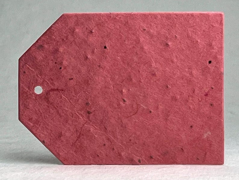 May include: A textured, tag-shaped label in a deep red hue. The tag has a small, circular hole near the top and a pointed bottom edge. The surface has a speckled appearance, suggesting it's made from recycled paper.