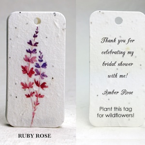 May include: A rectangular white paper tag with rounded corners and embedded dark flecks. One side features a watercolor illustration of a purple and red flower stalk. The other side displays the text: Thank you for celebrating my bridal shower with me! Amber Rose. Plant this tag for wildflowers!