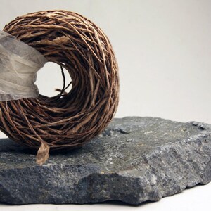 May include: A circular coil of brown fibrous twine secured with a strip of translucent white tape, resting on a jagged, dark gray piece of natural stone with a rough, uneven texture and visible mineral flecks throughout its surface.