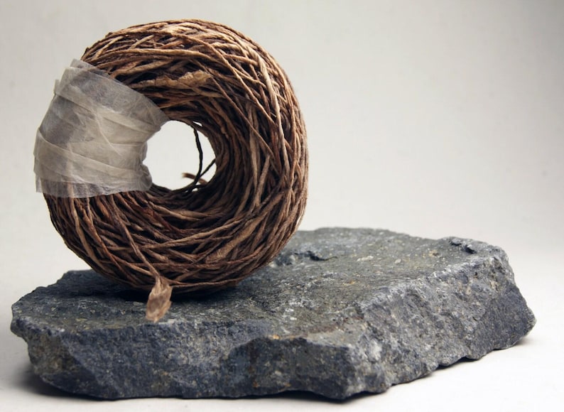 May include: A circular coil of brown fibrous twine secured with a strip of translucent white tape, resting on a jagged, dark gray piece of natural stone with a rough, uneven texture and visible mineral flecks throughout its surface.