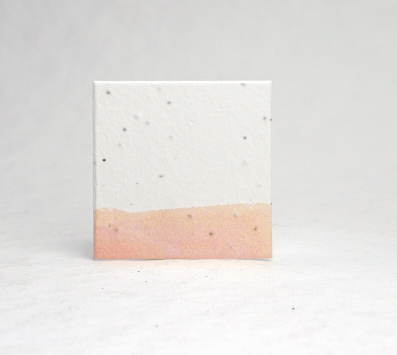 May include: A square card made of white paper with a light pink band across the bottom. The paper has small black flecks throughout.