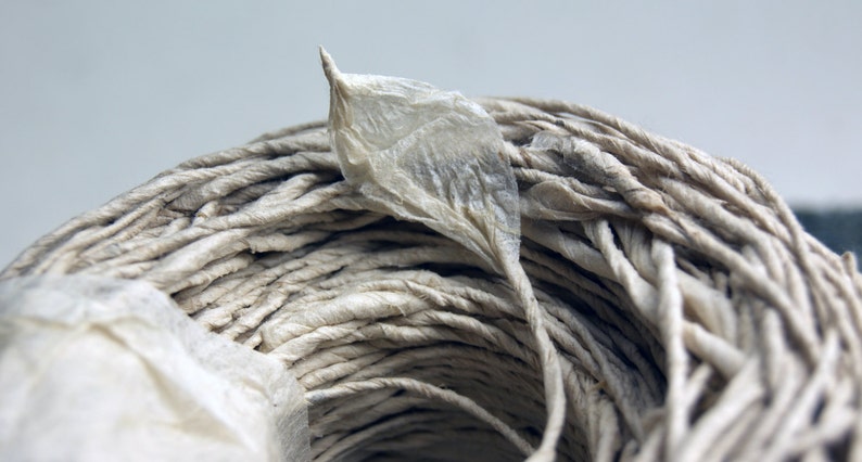 May include: A spool of light beige twisted paper twine featuring a single, delicate, translucent paper leaf attached to the top. The twine is wound into a circular coil, showcasing a textured, fibrous surface with a natural, neutral color.