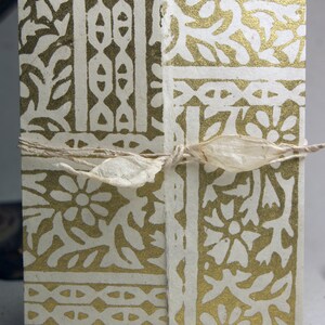 May include: A rectangular handmade paper journal featuring a metallic gold floral and geometric block print pattern on a cream-colored background. The journal is secured with a thin, natural-colored fibrous ribbon tied in a bow across the center.