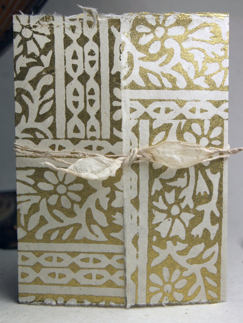 May include: A rectangular handmade paper journal featuring a metallic gold floral and geometric block print pattern on a cream-colored background. The journal is secured with a thin, natural-colored fibrous ribbon tied in a bow across the center.