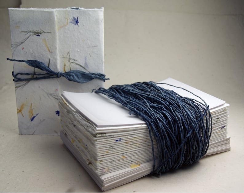 May include: Two handmade journals with blue string covers. The journal on the left is wrapped in a white paper with blue and yellow flowers. The journal on the right is wrapped in a white paper with blue string.