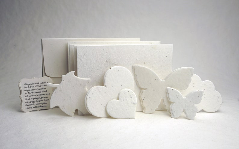 May include: A set of white paper cards and shapes made from recycled fibers. The paper is embedded with wildflower seeds. The text on the card reads: "This paper is made by hand from 100% tree-free recycled fibers. It contains our North American annual and perennial wildflower seed blend:  sapphire, petunia, daisy, poppy, forget-me-not, chamomile, cosmos, pink, calendula, and bear-tongue."