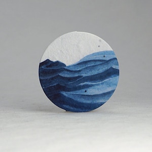 Pu&ograve; includere: Un sottobicchiere rotondo di carta bianca con un disegno ad acquerello di onde oceaniche in tonalit&agrave; blu. Le onde sono stratificate, creando profondit&agrave; e movimento. L'oggetto &egrave; decorativo.