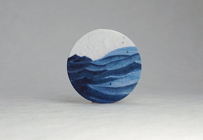Pu&ograve; includere: Un sottobicchiere rotondo di carta bianca con un disegno ad acquerello di onde oceaniche in tonalit&agrave; blu. Le onde sono stratificate, creando profondit&agrave; e movimento. L'oggetto &egrave; decorativo.