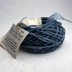 May include: A coil of blue paper twine labeled Eco-Twist in the color Spruce. The twine is wound into a circular shape and features a beige paper tag tied with a thin brown string. The tag includes printed text describing the product details.